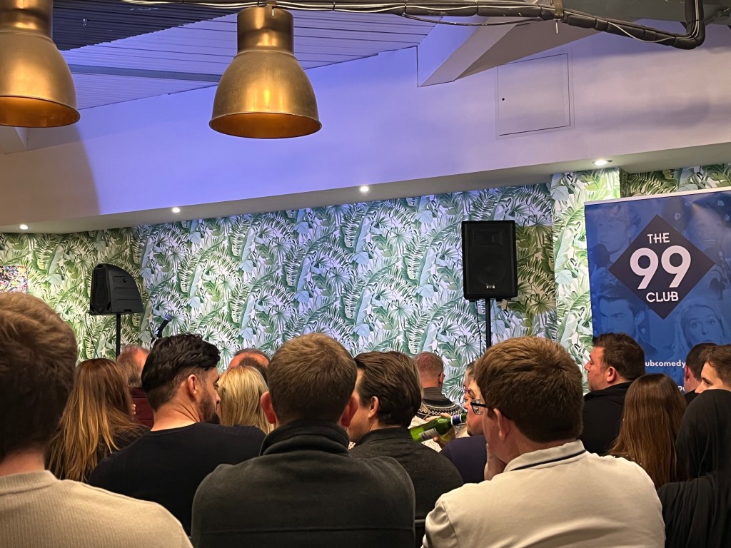 View towards the stage at the 99 Comedy Club in the Arboretum Leicester Square. There is a speaker on a tall stand on each side of the stage, while the back wall is white with large leafy designs on it. A stand to the right of the stage has The 99 Club in white text in a black diamond, which is placed upon a blue tinted background with pictures of comedians performing, including Josh Widdicombe and Jimmy Carr.