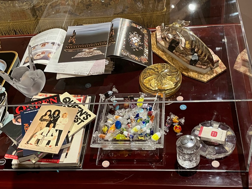 Various items on a table, including a collection of Sotheby's and Christie's auction catalogues, a collection of Queen books including songbooks, tour programmes and fan magazines, plus a selection of ornate glass items by Rene Lalique including boxes, a square dish containing colourful fake bonbon sweets, and a lighter with an ashtray.