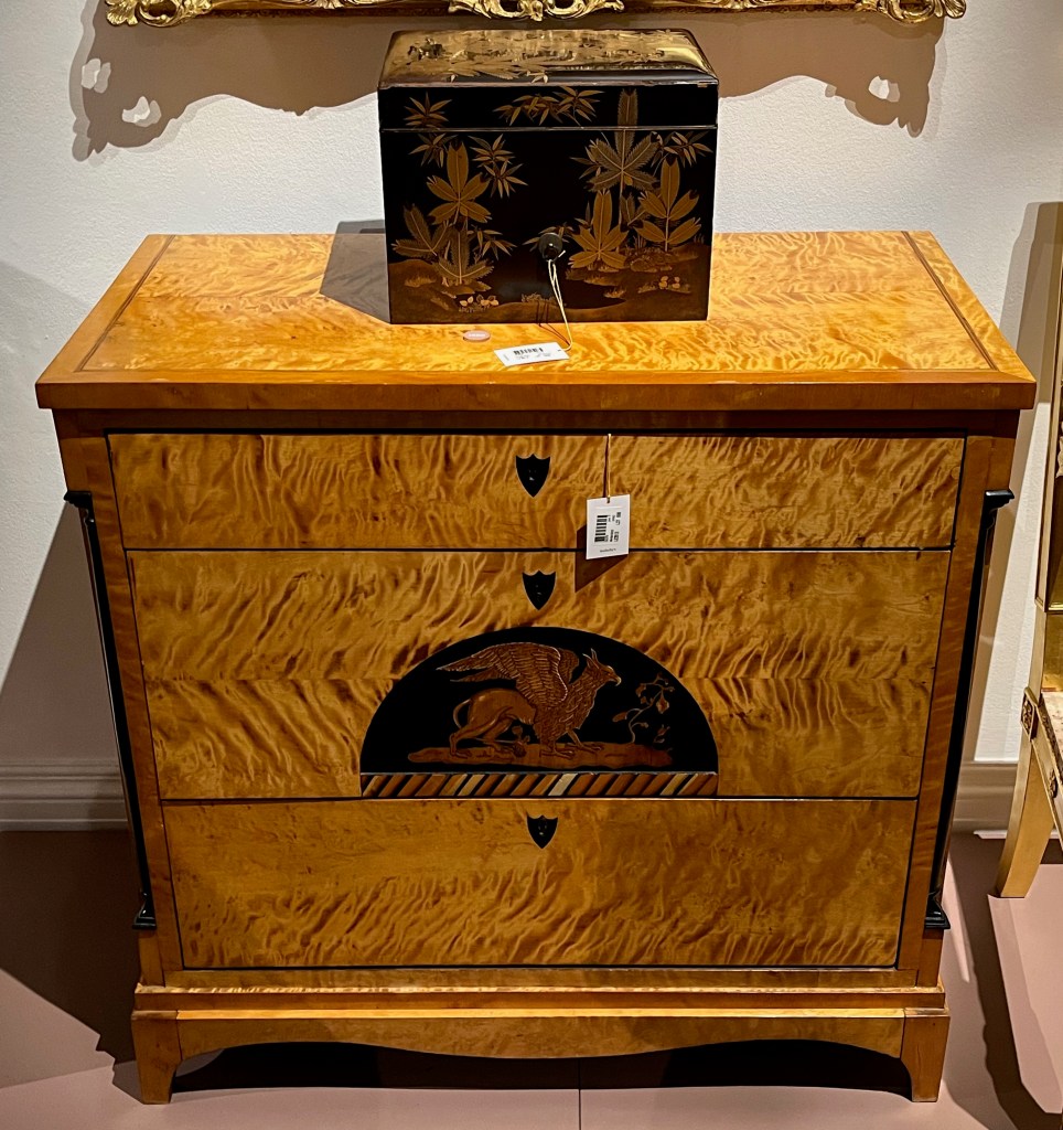 An ebony and maple chest of drawers, with one narrow drawer at the top and 2 larger ones beneath. The middle drawer has an arched painted panel in the centre depicting a griffin.