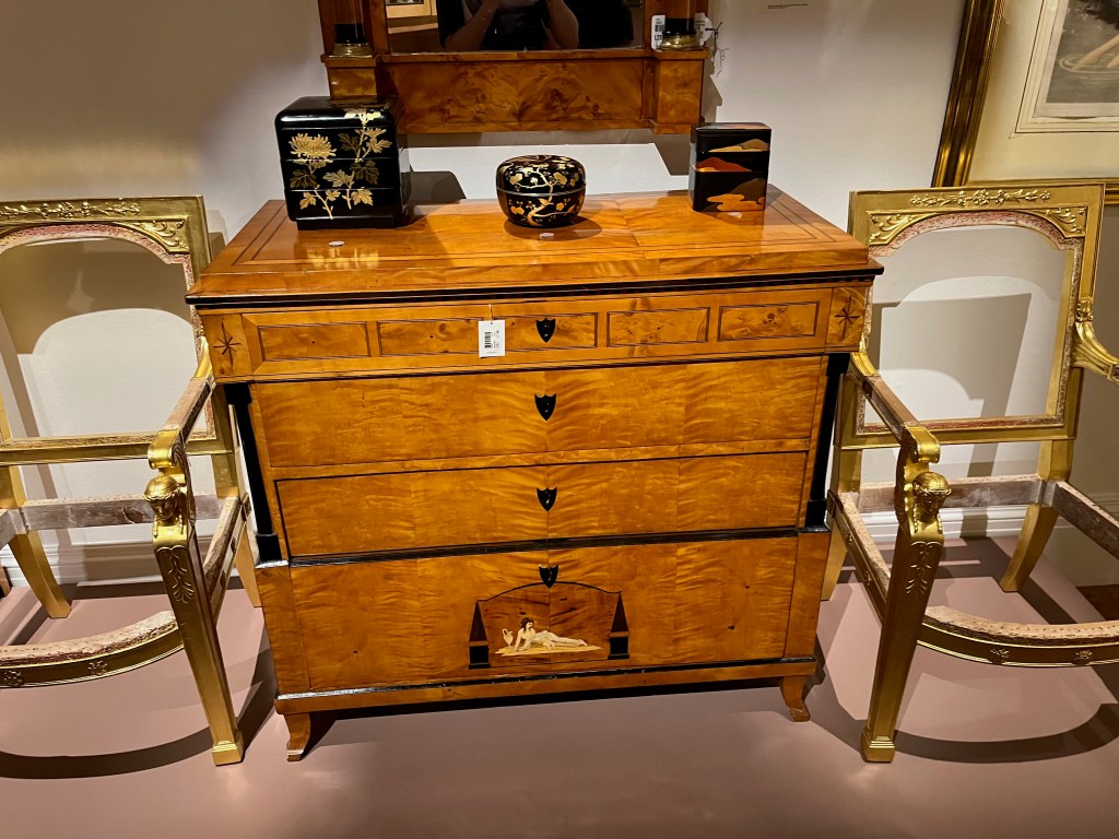 An ebony, parquetry and sycamore chest of drawers, with 1 narrow drawer at the top, 2 slightly larger drawers beneath, and a much larger drawer at the bottom. The bottom drawer has an arched painted panel in the centre of a lady stretching out on her side on the ground with a child sat next to her.