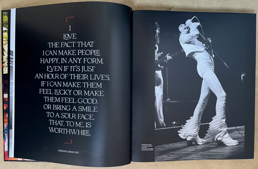 Double-page spread from the book for the Freddie Mercury exhibition at Sotheby's. On the right page is a black and white photo of Freddie, all dressed in white, leaning back as he sings into his microphone on stage. On the left page, in large white lettering on a black background, is a quote from Freddie that says I love the fact that I can make people happy, in any form Even if it's just an hour of their lives, if I can make them feel lucky or make them feel good, or bring a smile to a sour face, that, to me, is worthwhile.
