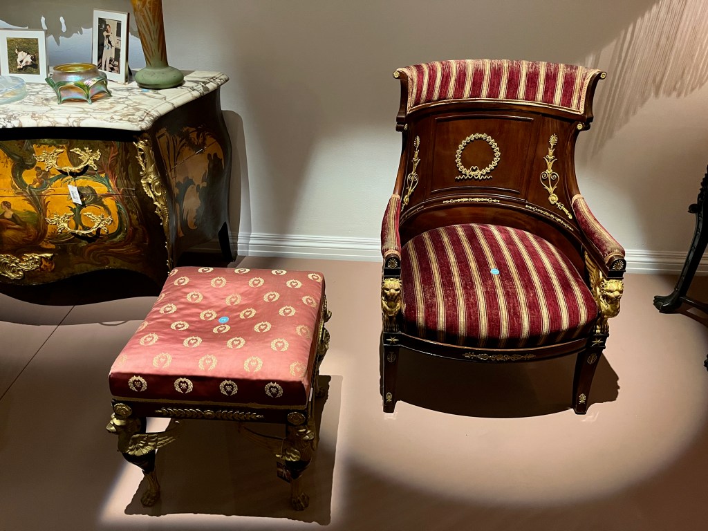 A mahogany stool with a padded seat upholstered in a red ground satin cover, with a design of repeating gold wing motifs. Next to this is a mahogany and giltwood armchair with red and cream striped velvet covers on the seat cushion and the top section of the back panel. Just beneath the front ends of the arms are gold supports in the form of winged lions.