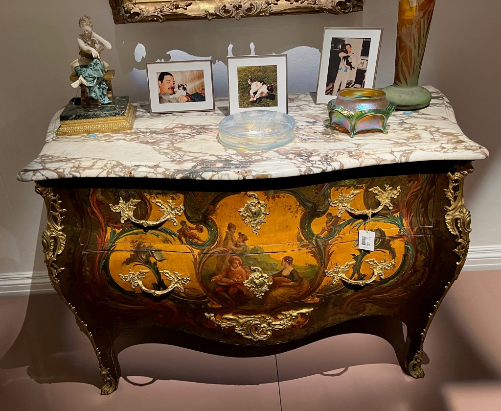 A French commode desk with a marble top on which are photos of Freddie Mercury and his cats. The front of the commode has 2 long drawers, one above the other, with ornate gold handles at each end, while the front overall is painted to show 4 people relaxing in a green landscape, along with other ornate detailing.