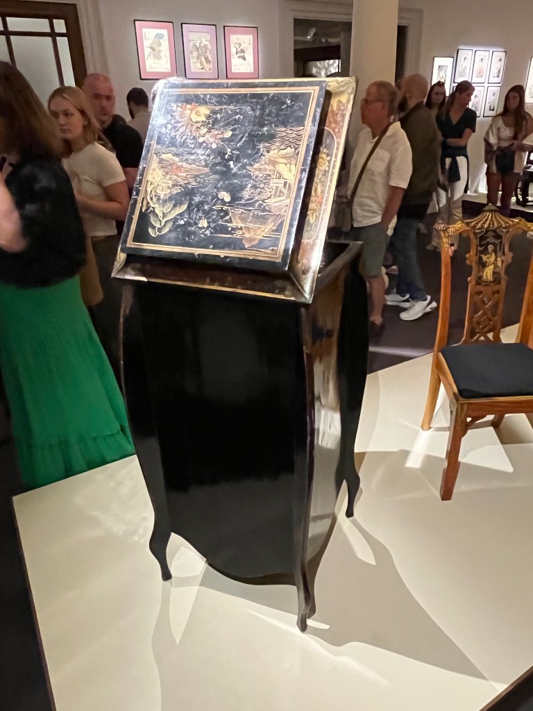 Rear view of a tall, free-standing cased gramophone. The back panel is plain, but the back of the lid at the top, which is open to reveal the record player at the front, is ornately decorated with gold leaf patterns.