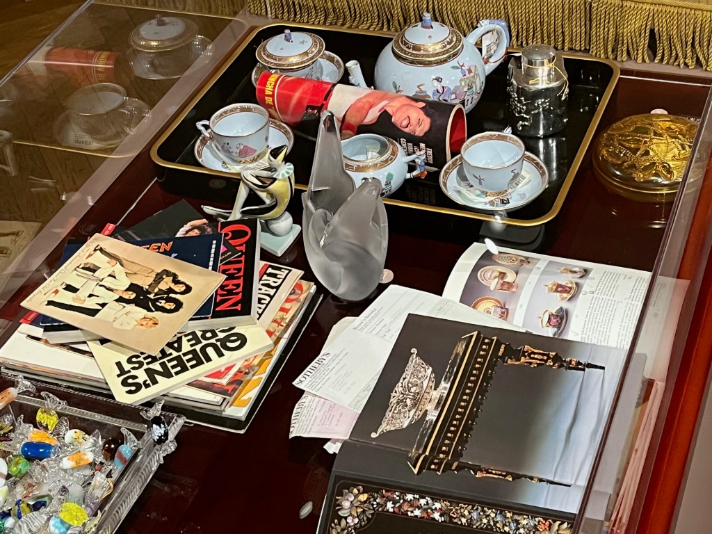 Various items on a table. At the top is a porcelain tea service, each piece painted with figures on a pale lavender background with gilded borders and pierced gilded collared feet. On the left is a collection of Queen books including songbooks, tour programmes and fan magazines. And on the right is a collection of Sotheby's and Christie's auction catalogues.