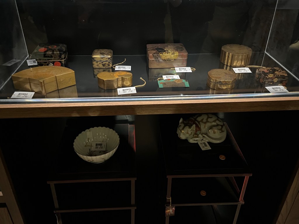 Two shelves of Japanese items. One item is a rectangular lacquer accessory box with rounded corners and overhanding cover, decorated in gold, silver, black and red with red fruiting vine.
There are also 4 rectangular incense boxes with flush-fitting covers, one decorated in gold and black depicting young pine, tree peony and oak leaf crests, another showing two court dancers beside a large drum, along with other musical instruments, a third decorated in gold, silver, black and red depicting a villa veranda beside a tree and grass fence, with butterflies among the foliage, and the 4th box shows blossoms and foliage, while the interior cover has maple leaves in autumnal colours. There's also a small comb decorated in gold, silver, black and red with a Dragon King mask among scattered maple leaves.