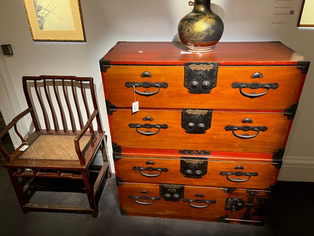 A large wooden Japanese chest of drawers on 4 levels. The top 3 levels have long drawers that span the width of the cabinet. But the bottom level is split, with a three-quarter length drawer on the left, and another compartment in the remaining space to its right. The wooden construction has a deep red tinge to it, and has ornate iron fittings, with 2 drop handles on the left and right of each drawer and an ornate decoration in the centre.