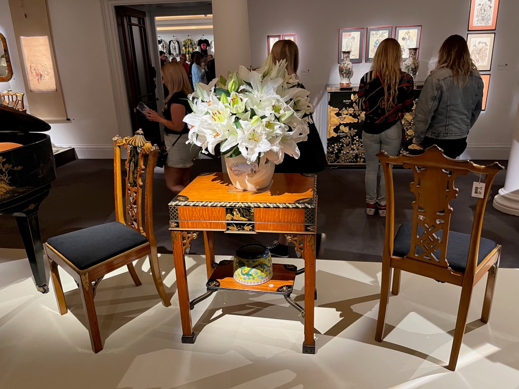 A square wooden table and 2 chairs. The table has an additional shelf underneath it, which is smaller than the top of the table, so is supported by poles diagonally coming out from each leg. A colourful bowl is on this lower shelf, while on the top of the table is a white vase with a scene painted on it that looks like someone by a rowing boat on a beach beneath a blue sky. The vase is filled with a huge, overflowing arrangement of white flowers. The back of each chair has an ornate carving going vertically up the centre, with space either side between the carving and the outer frame.