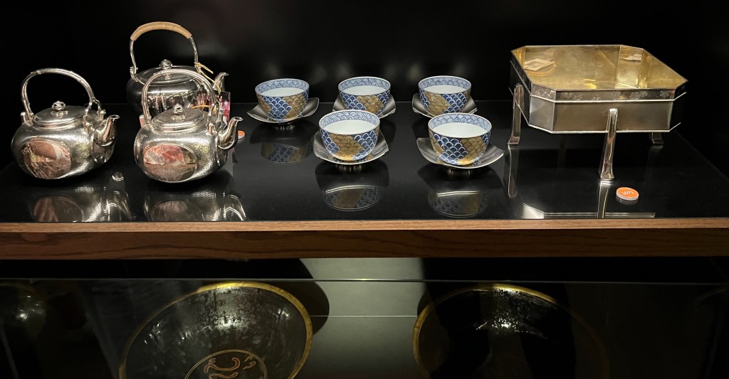 A Japanese tea set consisting of a silver ewer, five rectangular silver saucers that curve upwards at each end, and five cups decorated in coloured enamels on underglaze blue. To the left of the set is a pair of engraved silver ewers, inlaid in gold and silver with a central oval panel to the sides with chrysanthemums and bamboo. To the right of the tea set is a rectangular silver presentation box raised on four tall tapering feet, engraved to the rim with a band of stylised florets.