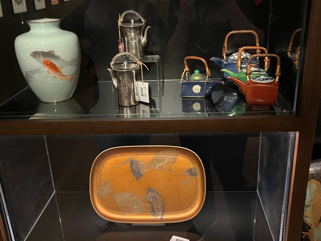 Two shelves with various Japanese items. On the top shelf is an enamel oviform vase with short neck, decorated in coloured enamels with carp swimming in still waters. Then there is a pair of silver ewers with tapering bodies, spout and woven sedge grass and silver wire loop handles. Next to those are four porcelain ewers in various sizes, decorated in blue, green and red enamels with gilt edges, each with a split woven bamboo loop handle. Then on the bottom shape is a rounded rectangular tray on four feet, decorated in gold and silver with fans, flowers and leaves.