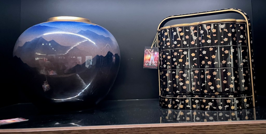 On the left is a large bulbous Japanese porcelain vase, decorated in various shades of underglaze blue and aubergine with mountains in mist, the shoulder applied with gold depicting a sunrise. On the right is a two-tiered box called a jubako, in the form of two overlapping blossoms with carrying frame, decorated in inlays of gold and silver foil on a black lacquer ground with scattered cherry blossoms.