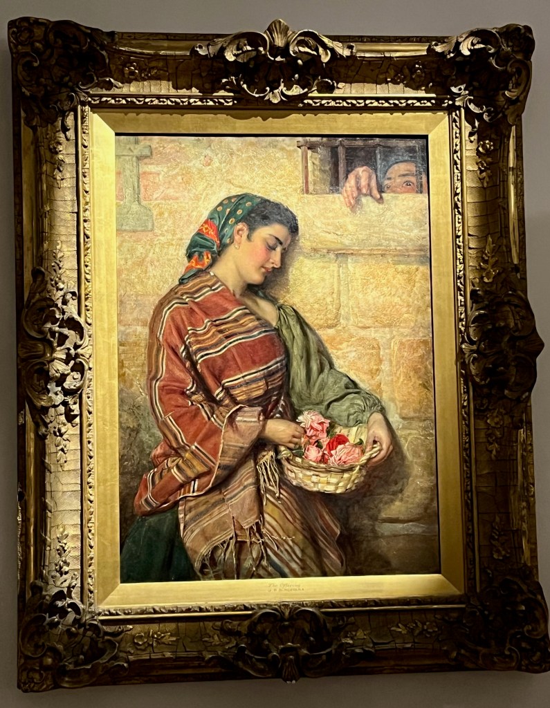 An oil on canvas painting by John Bagnold Burgess called The Offering. It depicts a woman wearing a colourful stripy shawl looking down at the small basket of pink flowers she's holding. She's standing next to a brick wall and just above her to her right is a small opening with a few vertical bars across it. Someone inside the room is reaching a hand through the bars towards the lady, but we can only see the top of their head down to their eyes.