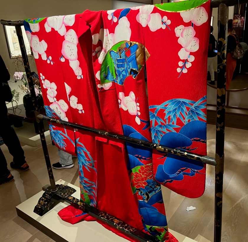 The back of a vibrantly coloured Japanese kimono. The thick long-sleeved robe is decorated in stencil dye with branches of blossoming plum, stalks of bamboo and pine interspersed with scattered fan-shaped images depicting ox-carts, peonies, pine, cranes in flight, chrysanthemums and irises.