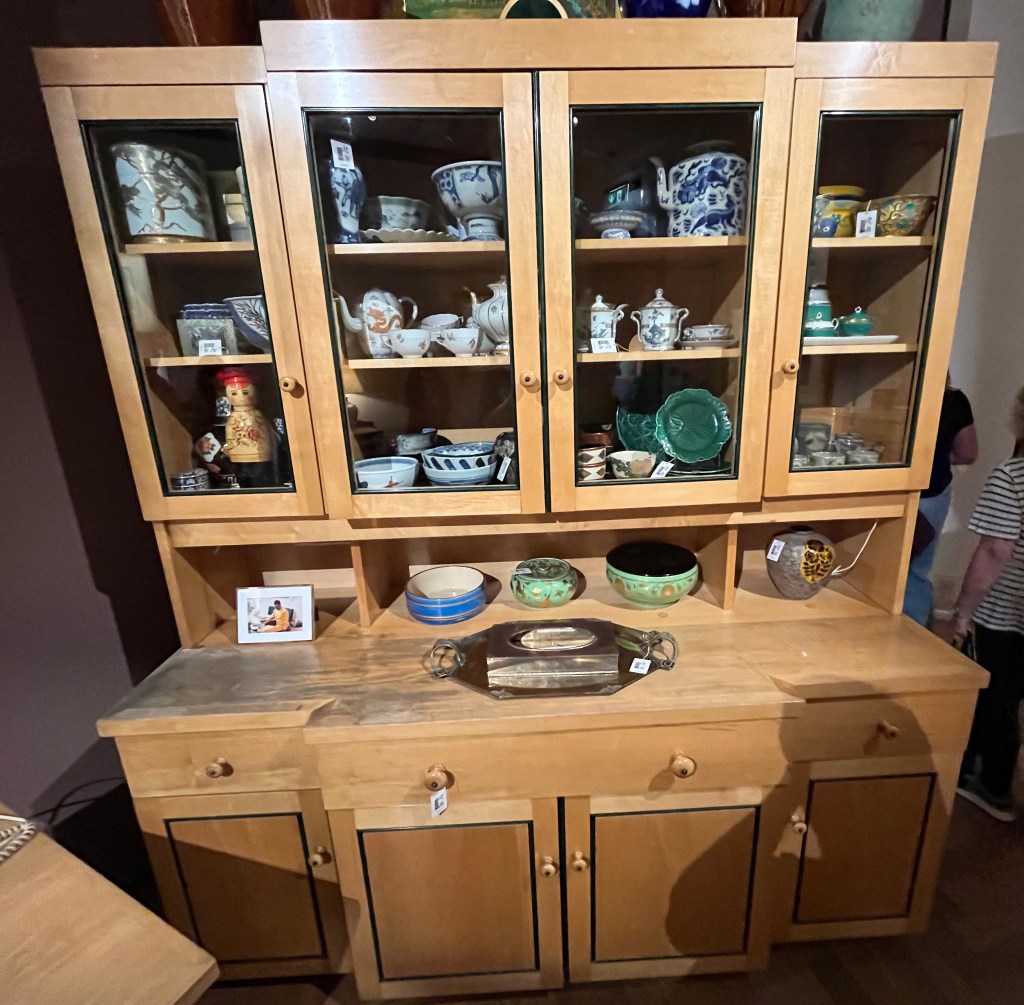 A large beech kitchen dresser. At the top are 2 single cupboards either side of a double cupboard, with clear glass windows so you can see the array of crockery inside. Below that is a large worktop area extending outwards, on which are a few bowls, a vase, a silver tray, and a small framed photo of Freddie Mercury wearing a yellow outfit, sitting down with what appears to be a black cat on his lap. Below the worktop are more cupboards, again with a single cupboard each side of a double. But this time they're just wooden doors with no glass, and there are drawers above them, single and double width to match the relevant cupboards.