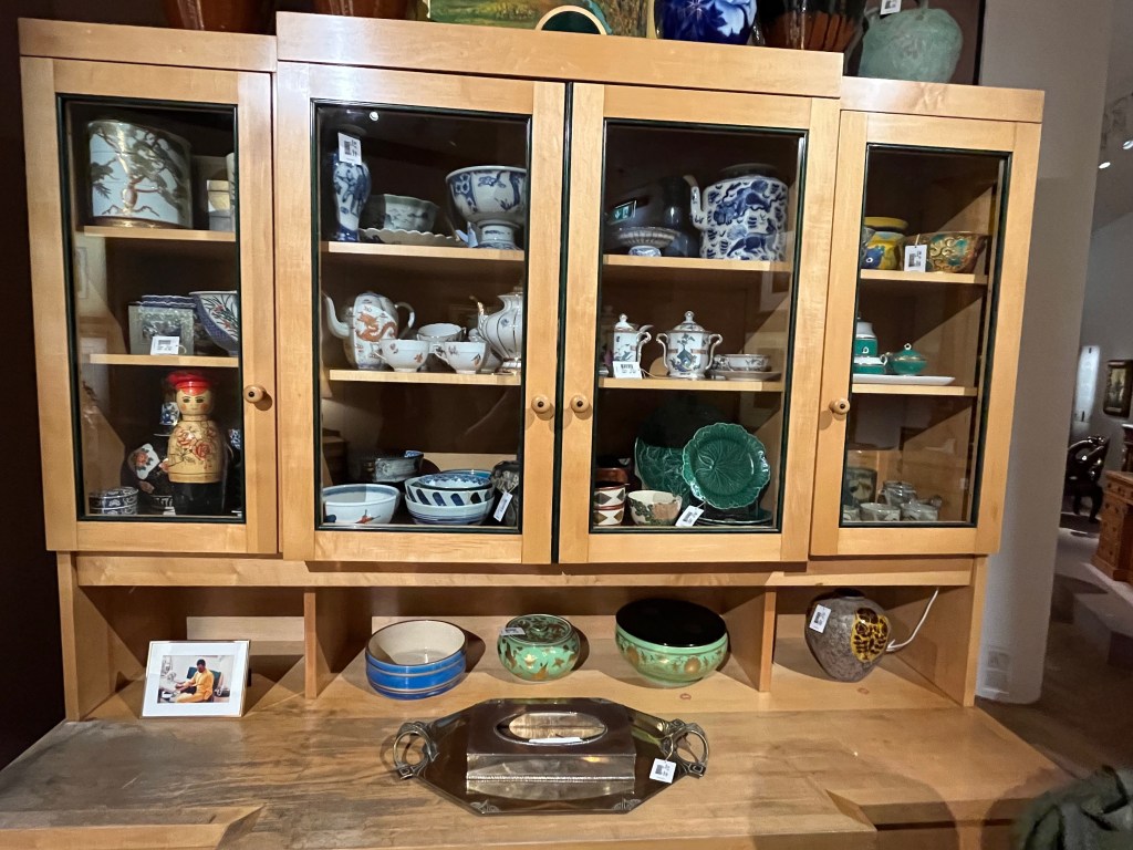 The top section of a large beech kitchen dresser. At the top are 2 single cupboards either side of a double cupboard, with clear glass windows so you can see the array of crockery inside. Below that is a large worktop area extending outwards, on which are a few bowls, a vase, a silver tray, and a small framed photo of Freddie Mercury wearing a yellow outfit, sitting down with what appears to be a black cat on his lap.