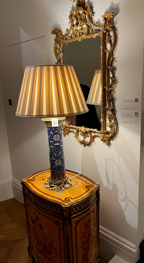 A lamp made from a tall, thin blue vase decorated with white flowery patterns, with a stripy lampshade. On the wall behind it as a George III mirror with a very elaborate and ornate gold frame.