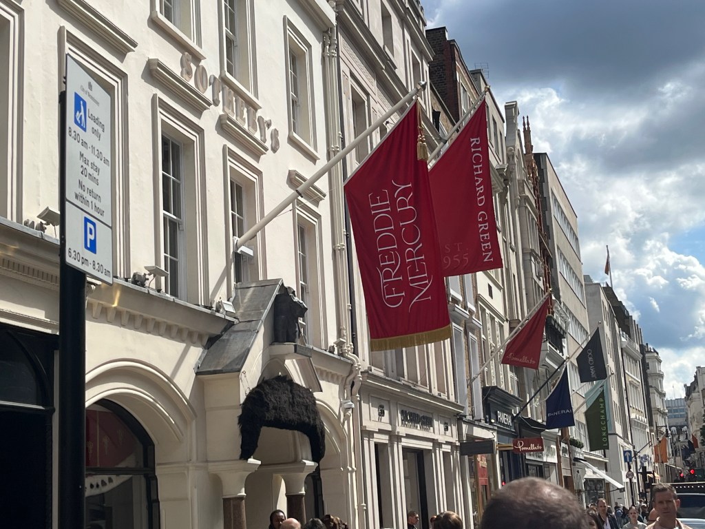The entrance to Sotheby's, with a large red flag hanging out from the building overhead, with large white text saying Freddie Mercury. There is some gold trim at the bottom of the flag.