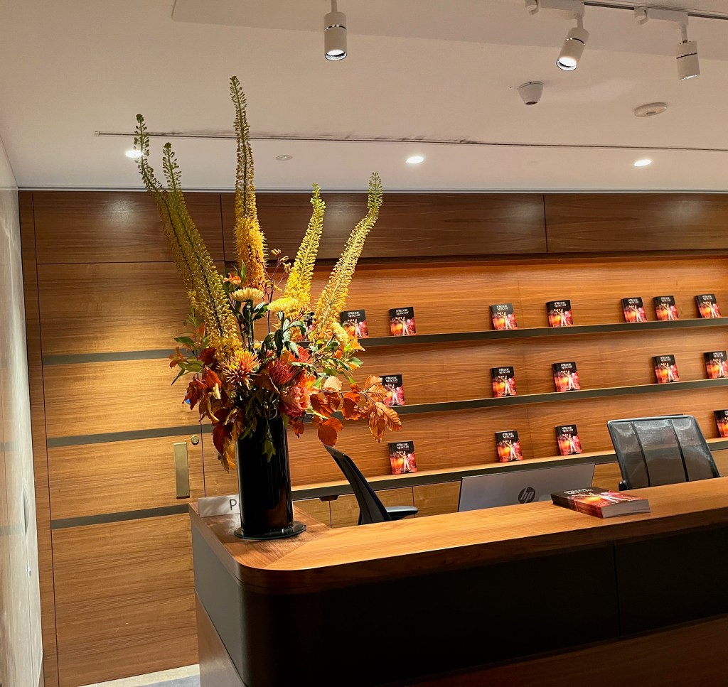 The reception in Sotheby's, with a huge arrangement of flowers at one end of the desk, while on the shelves at the back are copies of the book about the Freddie Mercury exhibition. One copy of the book is also on the desk, near the centre.