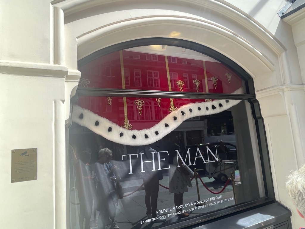 A large arched window, the top half of which is filled with the bottom of the regal robe worn by Freddie Mercury on tour, coloured red with gold decorations and white trim along the bottom. Below this, in the clear part of the window, is large white text simply saying The Man.