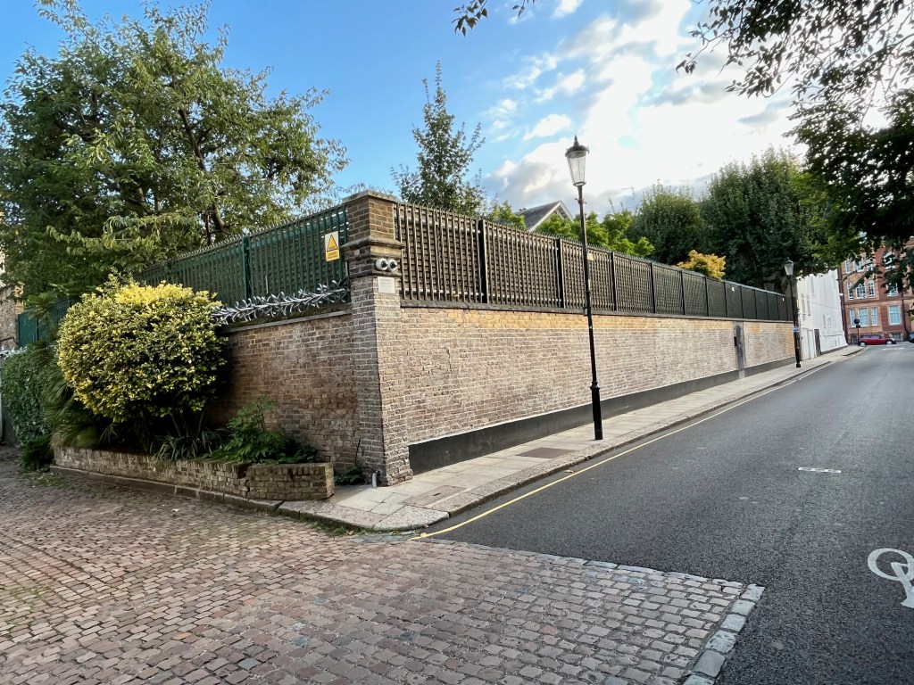The corner of the Garden Lodge estate, with the high brick wall topped by a large fence with razor wire. A small white sign attached on one side of the corner warns of CCTV in operation, while a small yellow sign just around the corner warns of the razor wire. A very large, round hedge is also growing by the wall around the corner.
