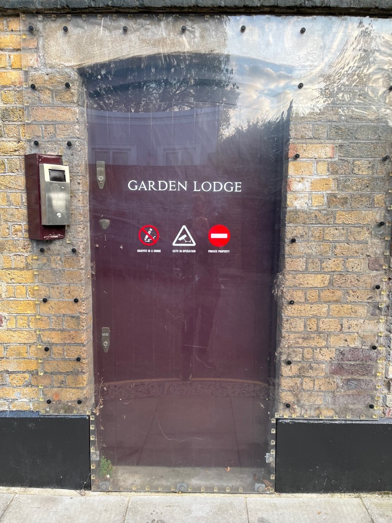 A black doorway in the wall of Garden Lodge, which has been completely covered by a large piece of solid plastic, which appears to be bolted to the brick wall around it. The plastic has white text saying Garden Lodge above 3 warning symbols. One says Graffiti Is A Crime below a white scribbling pen in a crossed out red circle. The second says CCTV In Operation with a white icon of a camera inside a white bordered triangle. And the third says Private Property below a no entry symbol, a red circle with a white bar across its centre. A metal intercom device is on the brick wall to the left of the door.