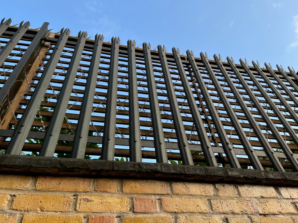 Close-up of the high slatted fence on top of the wall around Garden Lodge, through which you can see the razor wire on the other side.