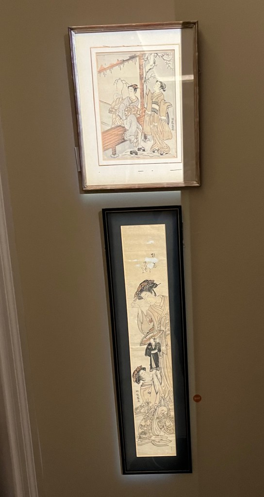2 Japanese woodblock prints by Isoda Koryusai. The first, in a regular portrait frame, shows a mother sitting on a veranda while her son plays with a branch. Snow has fallen and a young woman beside them breaks icicles from the eves with a stick. Below it is a very tall and thin artwork, in which the kamuro, or attendant for a courtesan, holds aloft a puppet modelled on popular kabuki figure Sukeroku, referred to as the Flower of Edo for his heroic exploits and bravado.