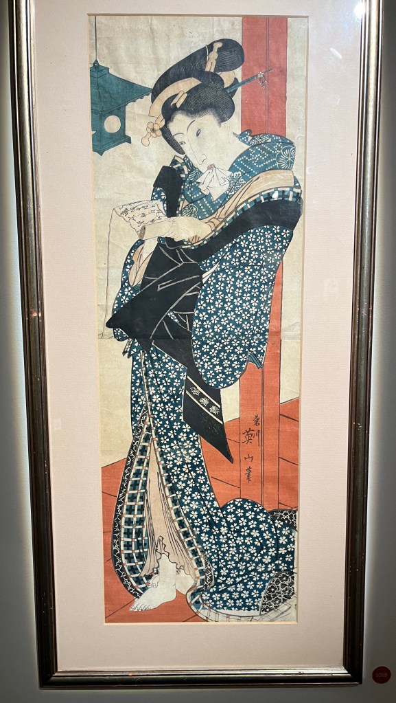 Japanese woodblock print by Kikugawa Eizan and Utagawa Kunisada. A lady dressed in a full length robe covered in lots of small white flowers reads a letter under the light of a lantern, while holding a small tissue in her mouth.
