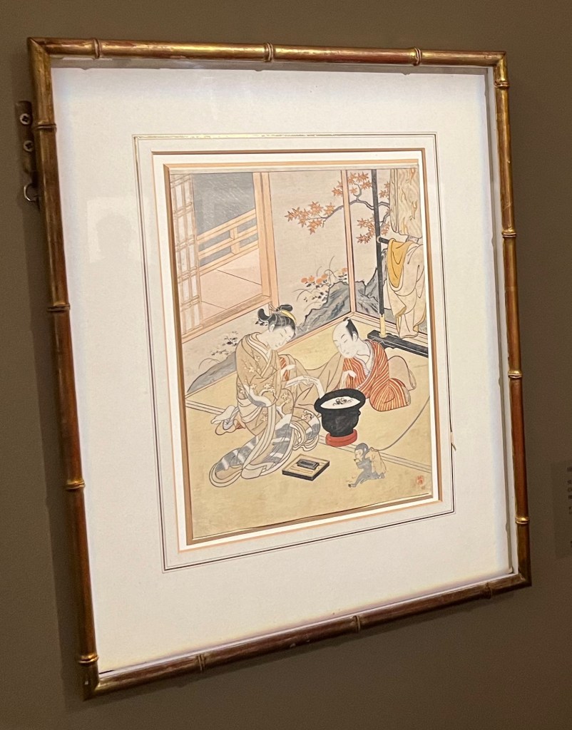 Japanese woodblock print by Suzuki Harunobu. On a cold evening in late autumn or early winter, a young courtesan dressed in a kimono with a design of snow-covered plum branches kneels beside a brazier whilst writing on a poem slip (called a tanzaku). Her client observes whilst reclining under winter robes. A tethered pet monkey sits near the brazier eating a fruit and further winter robes hang on a kimono stand (or iko) behind them.