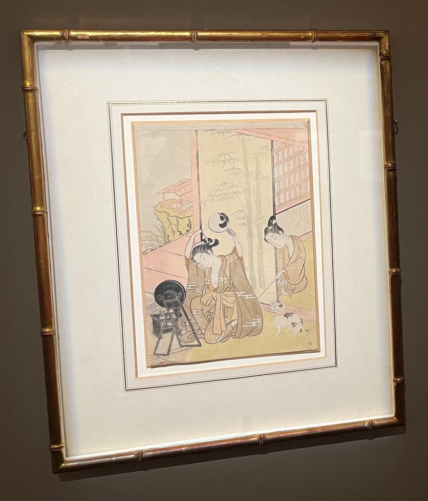 Japanese woodblock print by Suzuki Harunobu. A young woman in a room adjusts her hair using two mirrors, whilst her lover attempts to tickle her foot from behind a sliding door (shoji) with a stick. A cat appears about to pounce, excited by the movement of the stick.