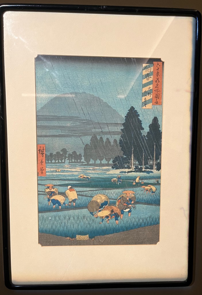 Japanese woodblock print by Utagawa Hiroshige, which shows people working on a field in the rain. A mountain looms high over the trees in the background.