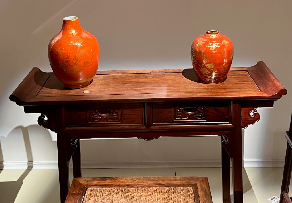 A Chinese hardwood console, the top curving slightly upwards at each end and 2 drawers beneath. On the table are 2 iron red vases that look bright orange in the light. Each is decorated in gilt on an iron red ground, the first has a tapering neck and has a design that depicts lotus leaves and flowers in meandering waters, while the second has a short neck and is decorated with autumnal flowers and grasses.