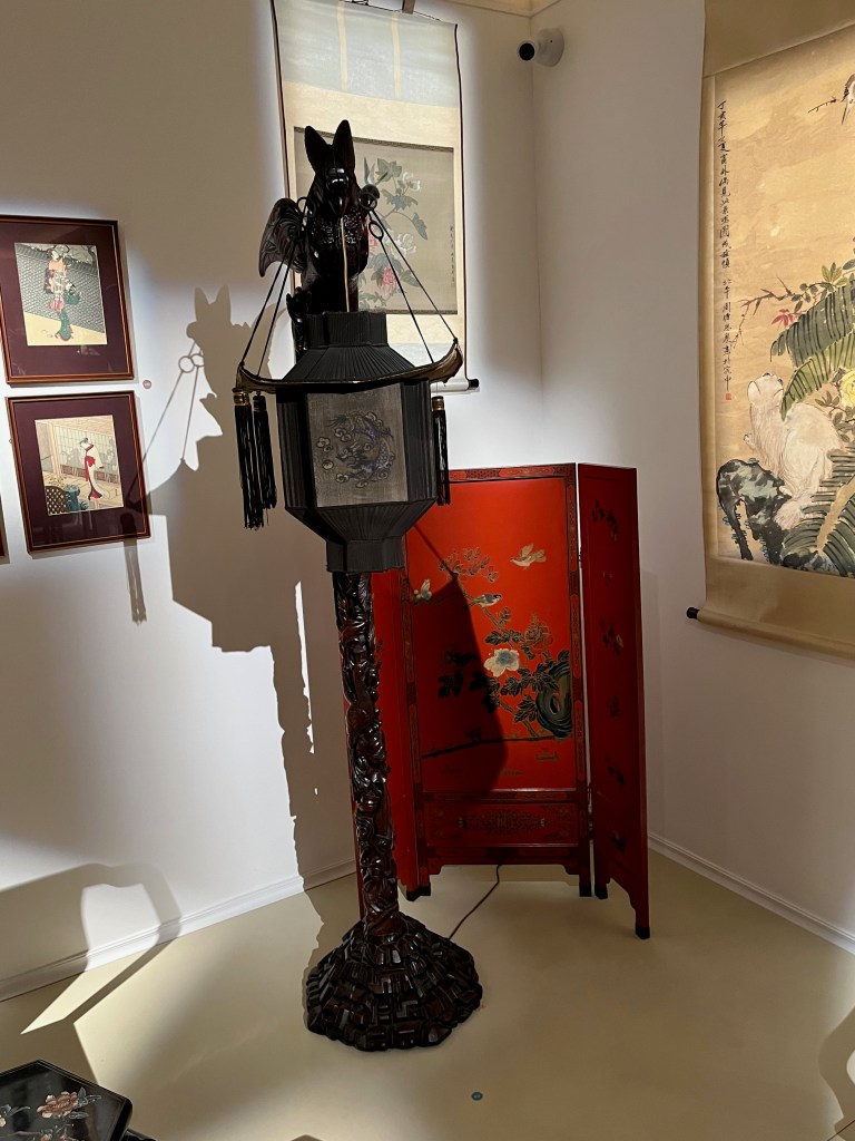 A Chinese lantern, black in colour, in the form of a winged phoenix on a tall fruiting and foliate stem and stepped rockwork base. In its beat, the phoenix is suspending a black and painted silk pagoda lantern.