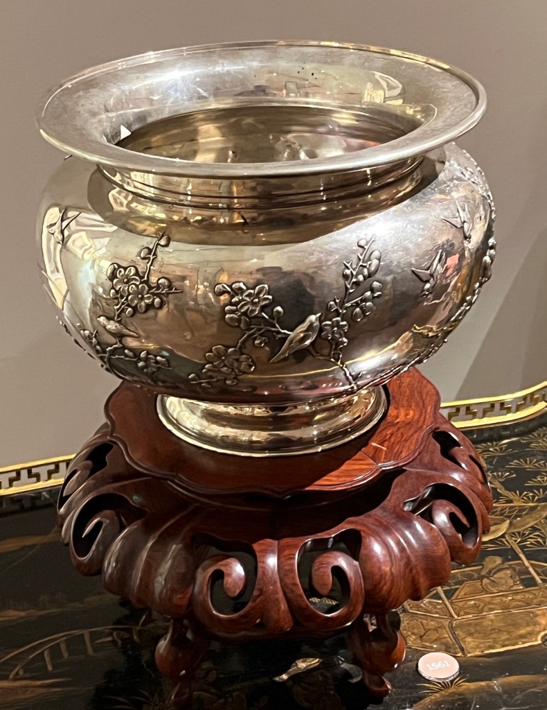 A large silver bowl called a jardiniere, with a bulging centre decorated with flowering branches and birds. It's stood on a round Chinese hardwood stand, with thick curly detailing carved around the edge.