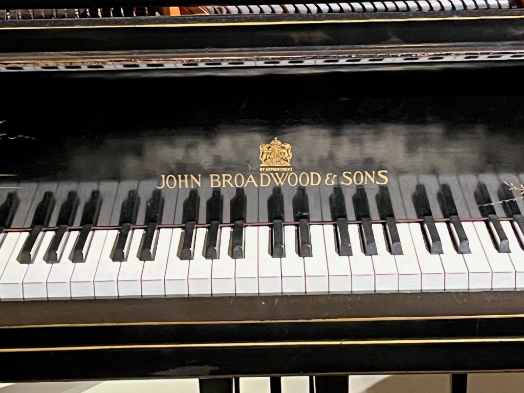Close up of the logo for John Broadwood & Sons, below a coat of arms and the words By Appointment, printed in gold above the keys on a black grand piano.