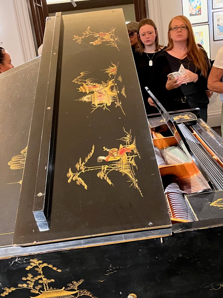 Decorations of stylised Japanese figures and buildings in predominantly gold colours on the raised top panel of a black grand piano.