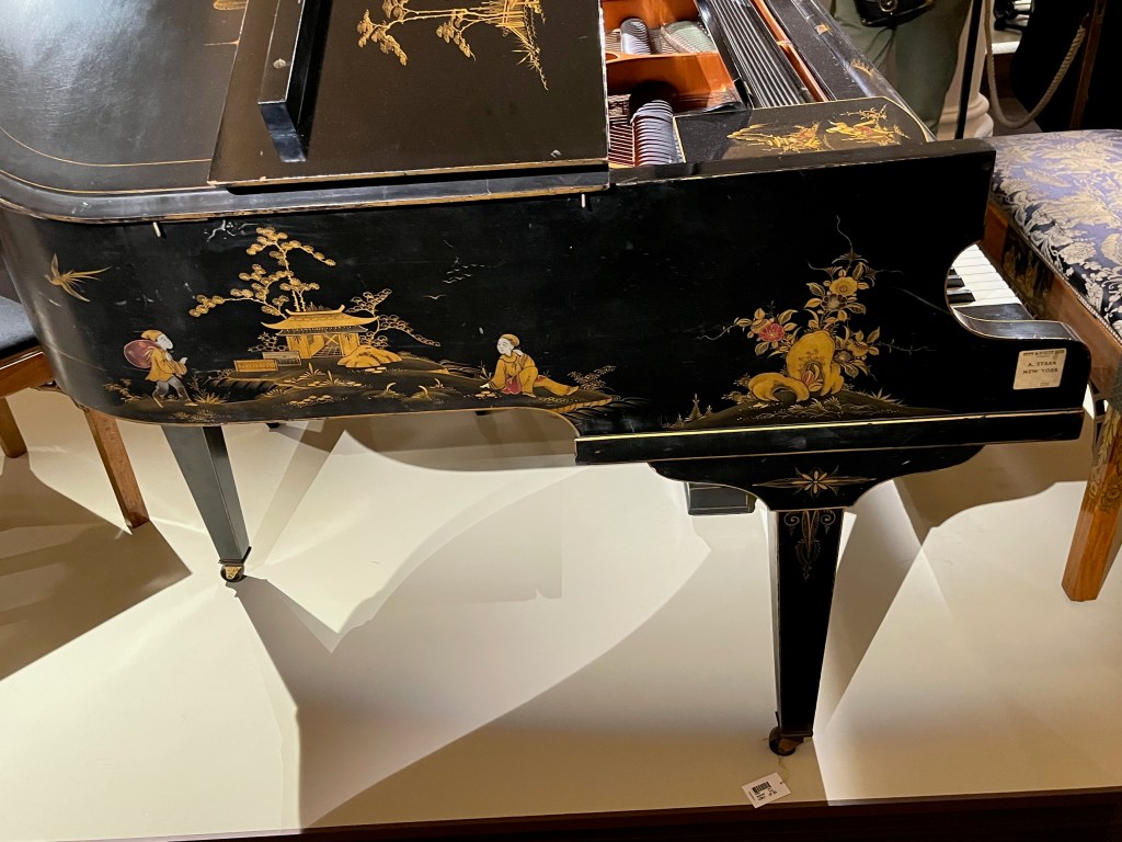 Decorations of stylised Japanese figures and buildings in predominantly gold colours on the left side panel of a black grand piano.