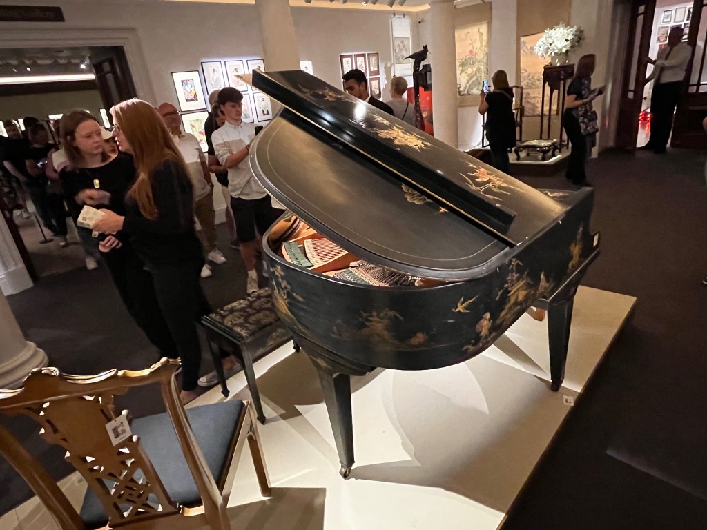 Rear view of a black grand piano decorated with figures in stylised landscapes, in the centre of a busy exhibition room in Sotheby's. The top panel of the piano is raised to reveal the strings and workings inside.