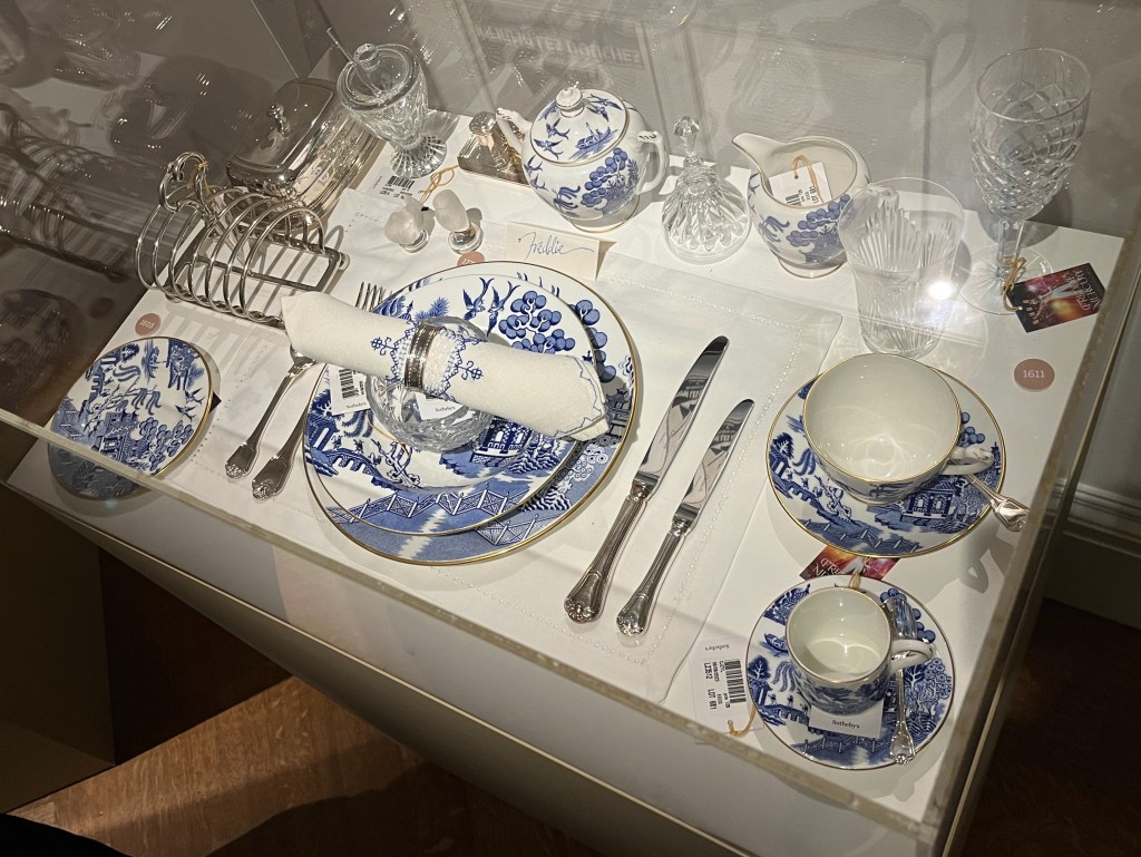 A dinner service with white plates and cups covered in artwork that uses shades of blue to create scenes of garden spaces, foliage and birds. Also on the table are some cut glass sherry glasses and a silver toast rack.