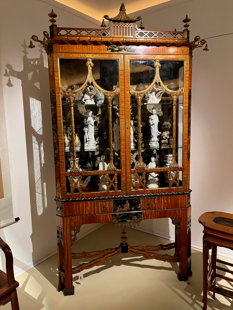 Tall wooden display cabinet featuring two glazed doors with ornate gold detailing curving across the windows, and a stylised pagoda on the top. The glass shelves inside are filled with little statues, vases and other trinkets.