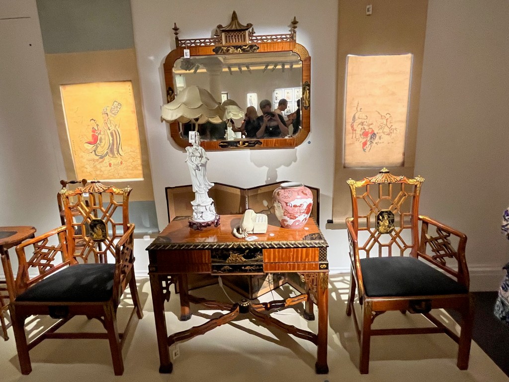 A wooden table with the beams between the legs connected in an X shape, next to two matching armchairs. A statue, push button phone and vase stand on the table. Above the table is a large, wide mirror, either side of which is a Japanese woodblock print.