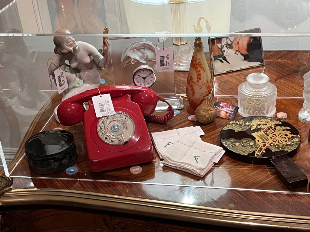 Various objects in a glass case on a table, including a red rotary telephone, an ornate black glass box, and a statue of a topless woman looking at herself in a handheld mirror.