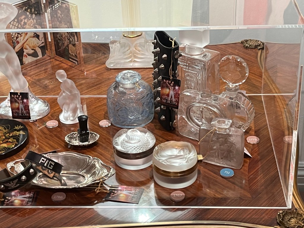 Various objects in a glass case on a table, including glass perfume bottles, an ink stamp and a small leather item with round metal spikes all the way down it.