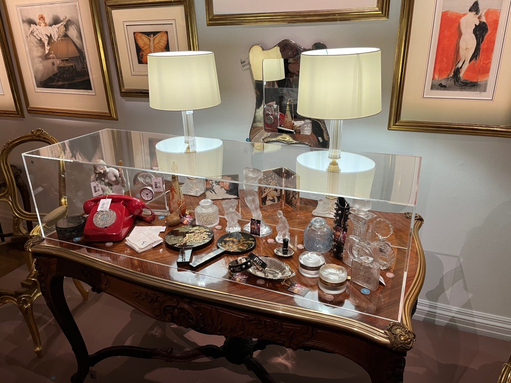 A French gilt-bronze mounted kingwood and carved walnut centre table, with wavy edges to the table top and some ornate leafy details around the sides of the table. The table is filled with a wide variety of objects, protected by a large glass case with 2 lamps standing on top of it.