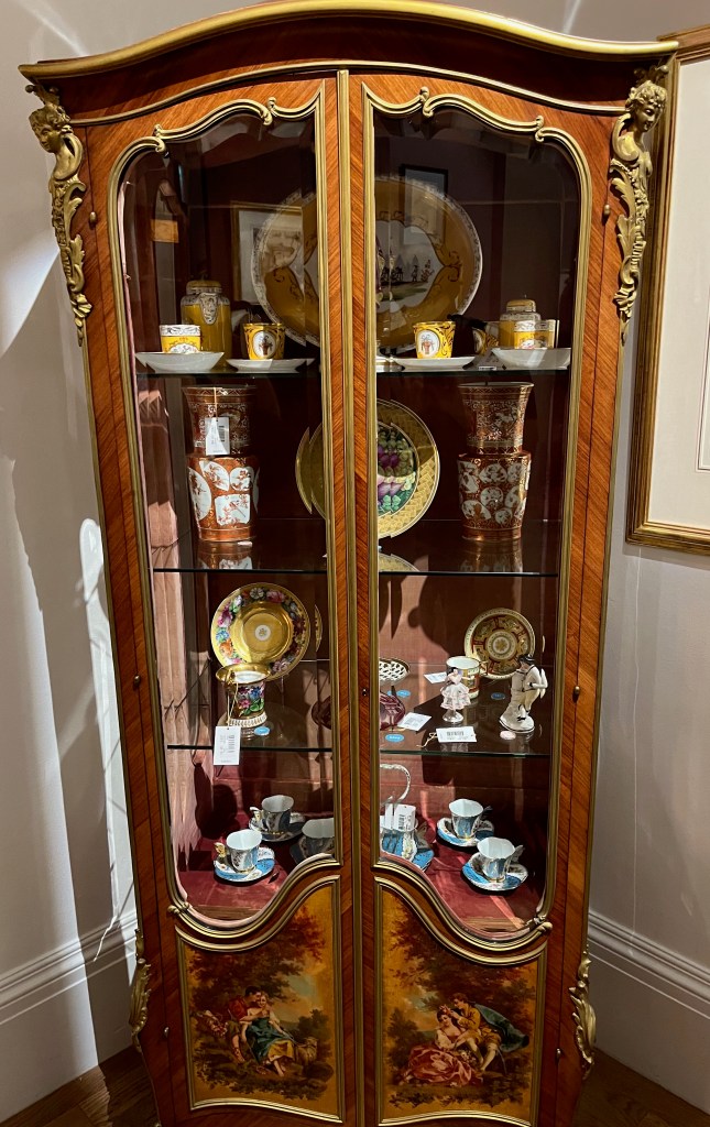 A tall gilt-bronze mounted 19th century display case with 2 doors featuring full length windows, curved edging and gold detailing. 3 glass shelves inside split the case into 4 sections, each of which is populated with colourful crockery and other items like little statues and vases.