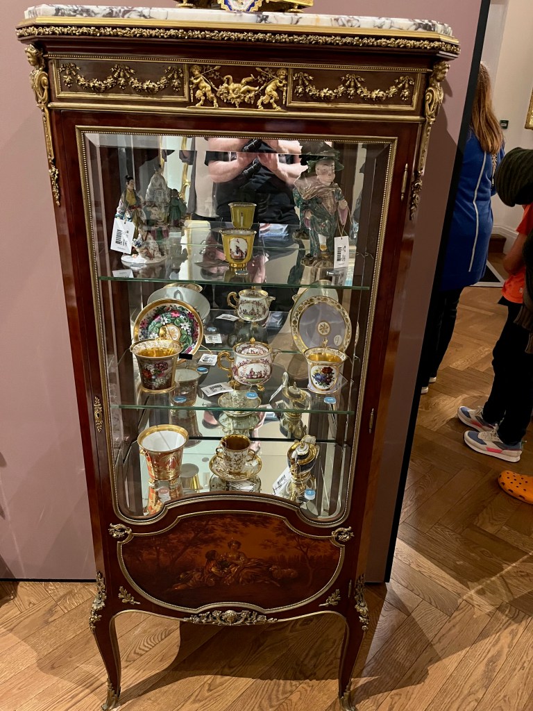 A wooden display cabinet with gold detailing across the top, and a painted panel at the bottom showing two seated figures and a sheep. Through the large glass window of the cabinet we can see the 2 glass shelves inside. These divide the cabinet into 3 spaces, which are filled with colourful crockery and statues. The back of the cabinet's interior is mirrored, reflecting its contents.