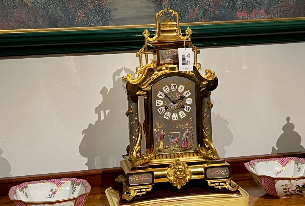 An ornate mantel clock with a gold frame, designed to look like a pagoda and decorated with figures amongst colourful flowering branches. The grey body has white patches with black Roman numerals forming the clock face.