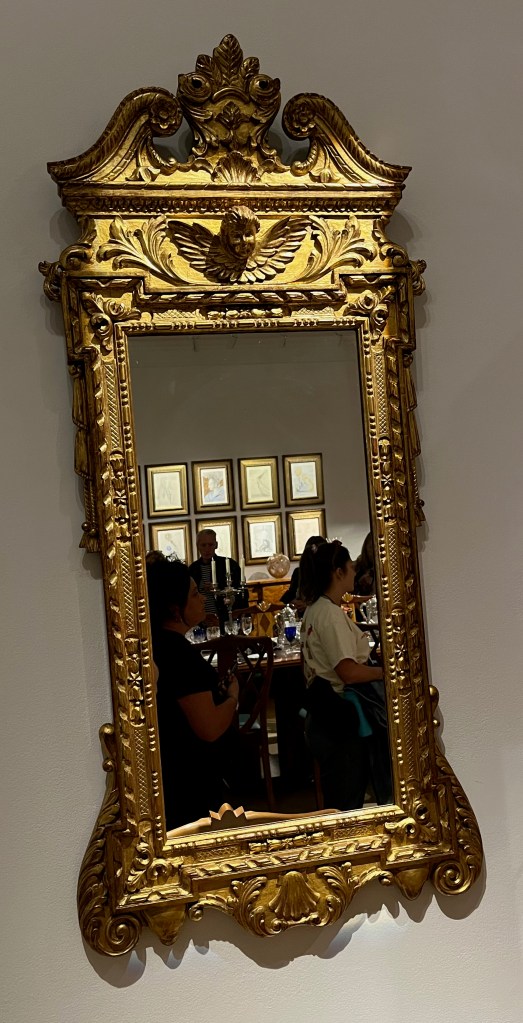 A George II style mirror with an ornately decorated gold frame, including foliage-style detail, and a small boy's head with wings either side at the top.