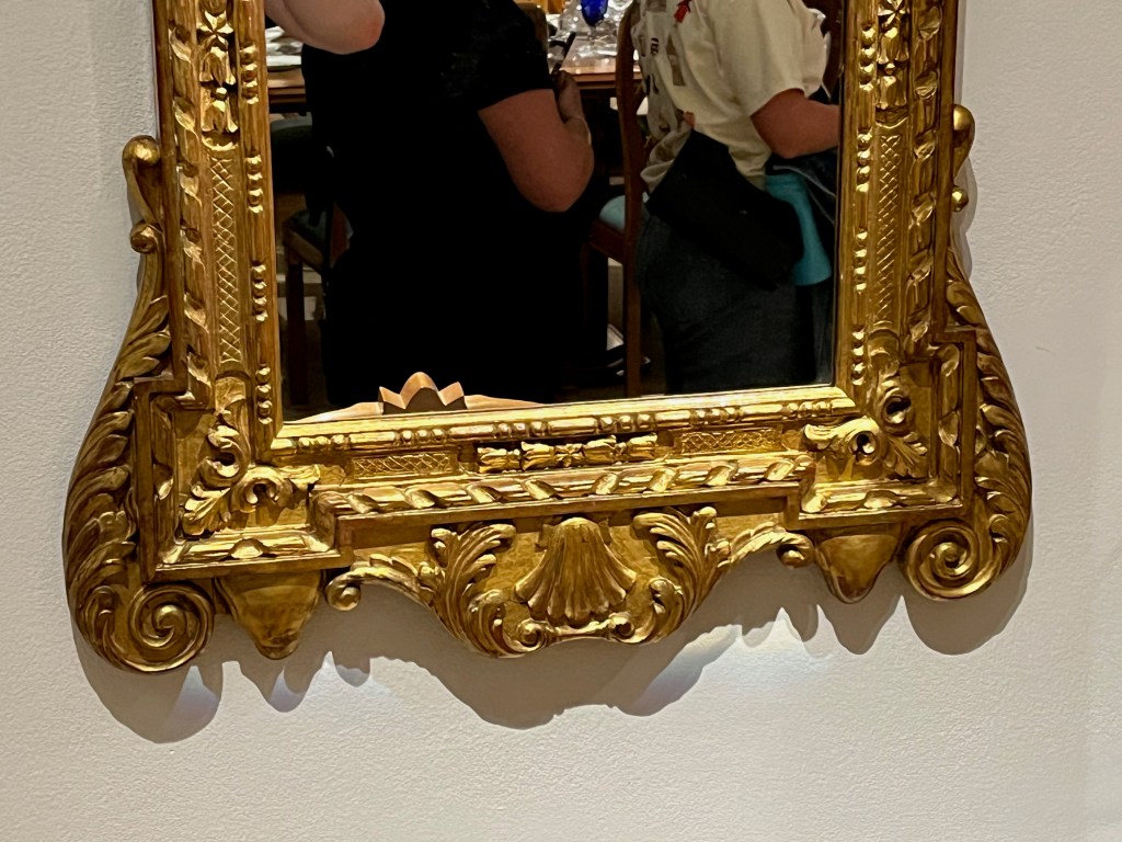 Close-up of the bottom of a George II style mirror with an ornately decorated gold frame, including foliage-style detail.