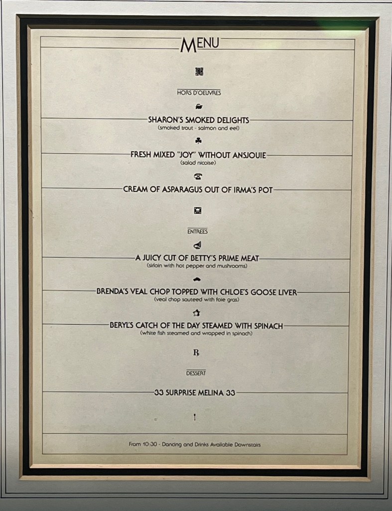 Black and white menu for the Infirma Les Douches charity event. Starters include Sharon's Smoked Delights, Fresh Mixed Joy Without Ansjouie and Cream of Asparagus out of Irma's Pot. Mains include A July Cut of Betty's Prime Meat, Brenda's Veal Chop topped with Chloe's Goose Liver and Beryl's Catch of the Day steamed with Spinach. Dessert is called 33 Surprise Melinda 33. A note at the bottom says from 10:30, dancing and drinks available downstairs.